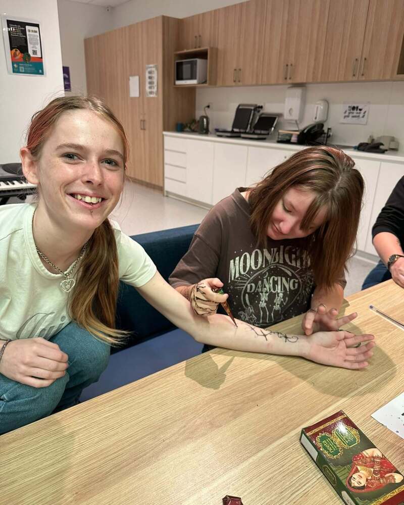 student smiling at camera whilst friend applies henna design to arm