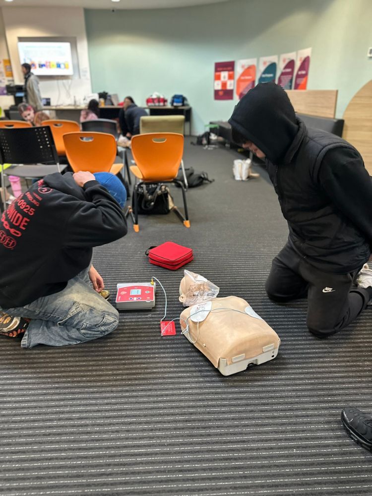 students in hoodies working with defibrilator on first aid dummy