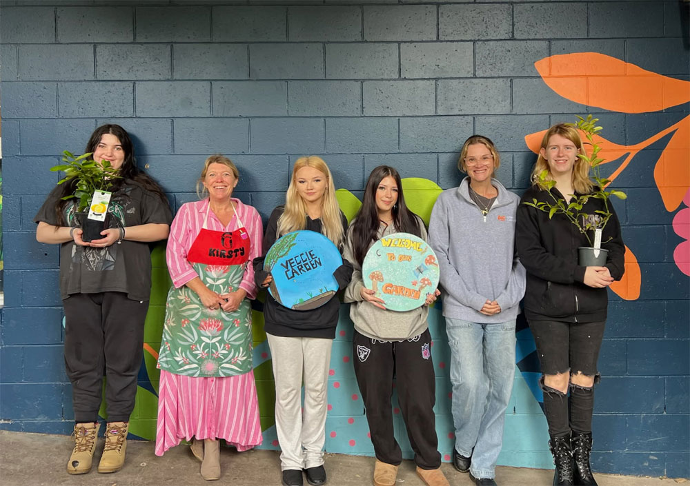 Students standing in front of a dark blue wall with veggie garden signs and plants in pots